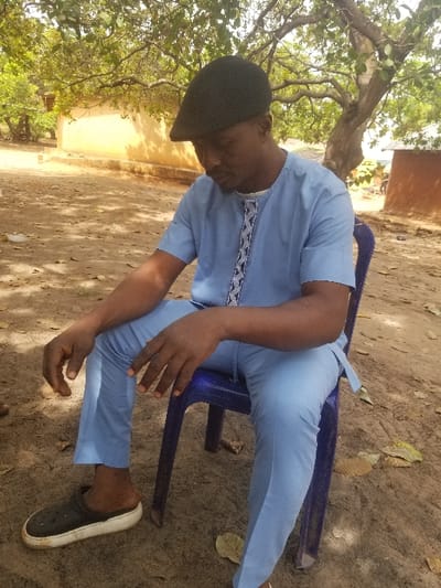 An outdoor scene in Ochadam, Nigeria, depicts an adult male seated on a blue plastic chair. The man, with dark skin, a beard, and a mustache, wears a dark flat cap, a light blue short-sleeved tunic with white and dark blue geometric embroidery down the front, and matching light blue trousers. His feet are clad in black and white slip-on clogs. He is looking downward and slightly to his right. His hands rest on his thighs. The ground is dry, reddish-brown earth with scattered light brown leaves. In the background, a large tree with visible branches and green foliage provides partial shade. Behind the tree, a single-story structure with light brown or yellowish walls is visible, suggesting an earthen or rendered building. The lighting indicates clear daylight.
