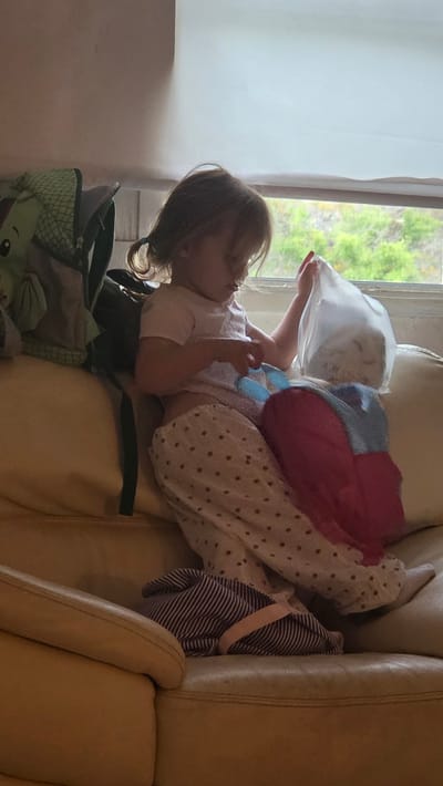 A young child is sitting on a tan sofa next to a window. The child is wearing a light pink shirt and patterned pants, and has their hair pulled back in two pigtails. They are holding a clear plastic bag in their right hand and are looking down at it. A pink and blue blanket is draped over their lap, and a striped bag is on the sofa to their left. A green backpack with a dinosaur design is on the sofa behind the child. The window shows a blurry view of green foliage and a cloudy sky. This scene is likely taking place in a residential setting in Guía de Isora, Spain, given the natural light suggesting daytime and the window view.
