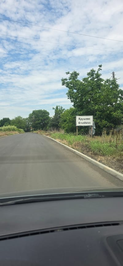 The image presents a forward-facing view from the interior of a vehicle, capturing an asphalt road leading into the distance. A white rectangular sign, supported by two dark posts, is prominently displayed on the right side of the road. The sign features text in black, identifying the location as "Крушево" (Cyrillic) and "Krushevo" (Latin script), indicating the entry point to the settlement of Krushevo, Bulgaria.
The road surface is dark asphalt, appearing dry, with a light-colored concrete curb delineating its right edge. Both sides of the road are flanked by dense green vegetation, primarily trees and tall grasses, which appear to be wild growth. The foliage is particularly thick on the right, partially obscuring the background behind the sign.
The sky occupies the upper portion of the frame, displaying a light blue color with numerous scattered white, fluffy clouds, indicative of daytime conditions. Two thin, dark utility wires are visible stretching horizontally across the upper sky. The dark-colored dashboard and the base of the windshield of the vehicle are visible in the lower foreground of the image. No individuals or animals are visible in the scene.