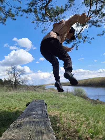The image captures a dynamic outdoor scene, with a person appearing to swing or hang from a tree branch against a bright blue sky. The photo is taken from a low angle, emphasizing the person's elevated position and movement.

The central subject is a person, dressed in a light beige or pale pink long-sleeved top and dark pants, with dark sneakers. They wear a dark cap or hat and over-ear headphones. Their left hand grips a pine tree branch, while their right arm is raised with fingers spread, contributing to a sense of playful motion.

The setting is a natural landscape, likely near a body of water. In the mid-ground, a wide river or similar waterway extends, bordered by a vibrant green, grassy bank. A bare-branched tree is visible in the distant background against the horizon. Closer to the foreground on the right, a thick, weathered wooden beam with prominent grain lies horizontally, possibly a part of a rustic bench, fence, or natural feature.

The time of day appears to be daytime, with clear, sunny weather indicated by the bright blue sky and scattered white cumulus clouds. The overall mood conveyed is one of freedom, activity, and enjoyment of nature. There is no visible text. The scene depicts a natural environment consistent with locations like Elets-Malanino, Russia.