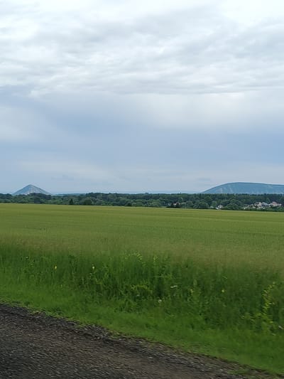 The image depicts a rural landscape with a vast green field in the foreground. In the background, a treeline separates the field from a distant town with houses. Beyond the town, two distinct mountain formations are visible on the horizon under a cloudy sky. The terrain appears to be generally flat with rolling hills and mountains in the distance. The image appears to be taken in the city of Vostochnyi, Russia, given its geographical context. The scene is peaceful and serene, suggesting a tranquil, natural environment.