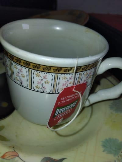 The image shows a close-up, slightly overhead view of a white teacup with a decorative band featuring a repeating pattern of brown squares and pink and yellow floral designs. A red tea tag, labeled "DANEDAR RICH, AROMATIC & REFRESHING TEA" in white and green text, hangs from the cup by a white string. The teacup sits on a saucer with a similar, faded floral pattern. The background is dark and out of focus, suggesting an indoor setting. There are no people or animals visible in the image. The scene is a still life focused on the teacup and its contents. The location is described as Chhina Uttar, Pakistan, but this information is not visually derivable from the image itself.