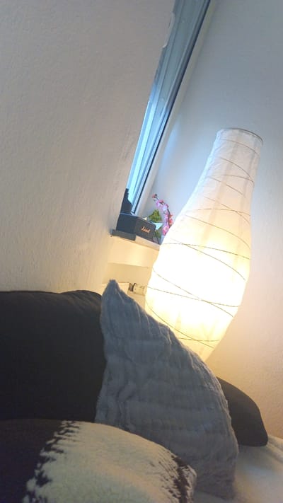A photograph depicts an interior scene, focusing on elements of a bedroom or living area. In the foreground is a stack of cushions/pillows, consisting of a dark-colored pillow, a blue pillow, and a white pillow with dark trim. The lighting casts a warm glow. Behind the pillows stands a tall, cylindrical lamp covered in a light, translucent material wrapped with string or wire. In the background, a windowsill is visible, containing a small black speaker, possibly a Marshall product, and a small plant with pink flowers. The windowsill is positioned in front of a window angled into the wall. The photograph was taken indoors, potentially in a residence located in the city of Ginsheim-Gustavsburg, Germany.