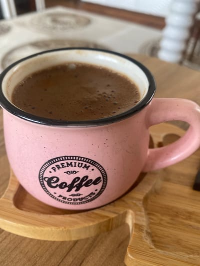 A close-up shot shows a pink ceramic mug filled with dark coffee, resting on a light brown wooden tray. The mug has a black rim and a black circular logo on its side that reads "PREMIUM COFFEE PRODUCTS" in a stylized font. The coffee has a frothy top. The wooden tray has a textured surface and is positioned on a wooden table. In the blurred background, a white tablecloth with a circular pattern is visible, and a portion of a wooden chair can also be seen. The overall scene suggests a relaxed setting, likely a home or cafe, in Talas, Turkey.