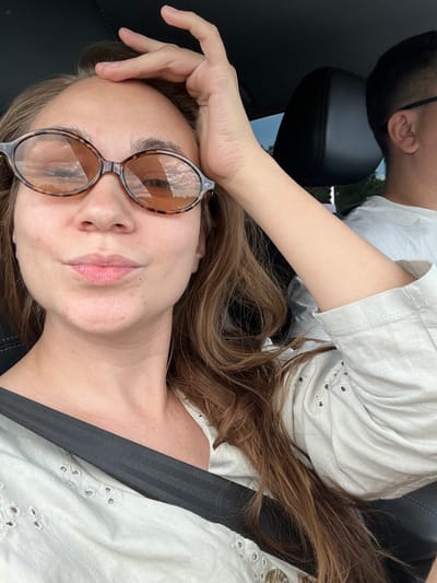 A woman with auburn hair is in the passenger seat of a car, looking at the camera. She is wearing a light-colored top with intricate cutwork detailing around the neckline and sleeves, and a dark seatbelt across her chest. Large, tortoiseshell-patterned sunglasses with amber lenses obscure her eyes, and her left hand is raised to her forehead. The reflections in her sunglasses show a sky with clouds. To her right, a man with short dark hair and glasses is visible in the driver's seat. The interior of the car appears to be dark, with a black leather headrest visible behind the woman. The overall scene suggests a casual journey, possibly during daylight hours given the lighting and reflections. The image does not provide specific visual cues to indicate a location within the city of Pervomaiskoe, Russia.