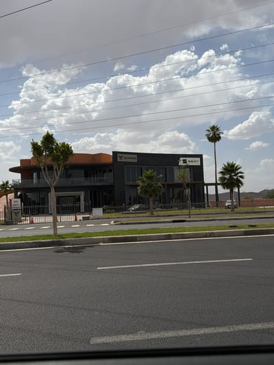 The image depicts a modern, dark-colored building with signage indicating it's a SEAT and CUPRA dealership, located in Ouahat Sidi Brahim, Morocco. The building is situated next to a road with white lane markings and a grassy median. Palm trees are planted near the building. The sky is cloudy, with power lines visible against the sky.