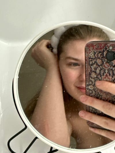 A selfie captured in a round mirror, reflecting a person's face and arm. The person is holding a phone with a patterned case, partially obscuring their face. Their expression is somewhat somber. The mirror sits on a white surface with a black wire rack visible in the background. The lighting is soft, creating subtle shadows. The reflection reveals a close-up perspective, focusing on the face and upper body.