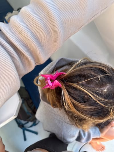 An overhead view captures a partial scene inside an enclosed space, identified as the interior cabin of a boat. The central subject is the upper body of an individual, primarily focused on the back of their head and shoulders. This person has medium-brown hair with blonde highlights, partially secured by a bright fuchsia-pink, five-pointed claw-style hair clip. They are wearing a light gray hooded sweatshirt with the hood up. A ribbed, light beige or off-white long-sleeved garment covers an arm extending from the upper left of the frame. In the lower right, a bare foot with light-colored toenails rests on a clean, light-colored floor surface. The background features a dark blue upholstered surface behind the individual's left shoulder, with a dark bag and a blue strap visible below it. A plain white vertical panel forms the background to the right. The scene is illuminated by natural light.