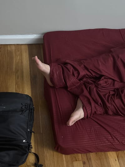 An overhead shot captures a person resting or sleeping on a maroon bed in an indoor setting. The person's bare feet extend from under crumpled maroon bedding or clothes, which cover the majority of their body and the bed. The maroon fabric exhibits a subtle striped texture. To the left, a light grey wall meets the wooden floor. The floor, composed of light-toned wooden planks, fills the lower and right portions of the frame. In the lower right corner, a black duffel bag or backpack with visible zippers and straps rests on the wooden floor. The scene suggests a private room, possibly a bedroom, within a dwelling in Franklin Township, United States of America. No activity beyond resting is discernible, and no text is visible.