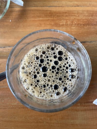 A top-down, close-up shot captures a glass mug filled with dark coffee topped with a thick layer of foam. The foam is a mix of small and large bubbles, with some of the larger bubbles appearing dark and glossy. The coffee is set on a wooden table with visible grain patterns. In the upper left and right corners, portions of what appear to be white paper napkins or food wrappers are visible. The image is well-lit, with natural light casting a soft glow on the scene. The specific location in Pulokiton, Indonesia is not discernable from the visual information provided.