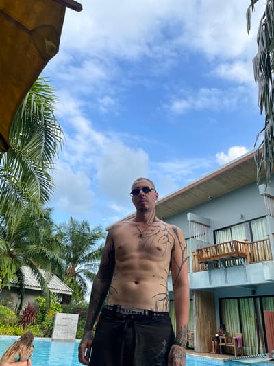 This image depicts an outdoor scene in the City of Ban Tha Muang, Thailand. A male individual stands centrally in the foreground, facing the camera. He has short, light-colored hair, wears dark sunglasses, and a dark pendant on a chain around his neck. His body is extensively tattooed with black ink, including full sleeves on both arms and abstract, tree-like patterns covering his chest, torso, and extending up his neck. He wears dark swim shorts, with a visible grey waistband bearing the "LUPO" logo.
Behind the male subject is a swimming pool. To the lower left, a second individual with long, light-brown hair, wearing a turquoise bikini top, is partially visible at the edge of the pool. In the background, a light-blue multi-story building, likely a hotel or guesthouse, features a wooden balcony on its second floor and visible windows. A third individual is seated on a wooden bench on the ground-level patio of this building, appearing to interact with a handheld device. The environment is rich with green palm trees and other tropical foliage, under a clear blue sky with scattered white clouds. A partial view of a yellow fabric awning is visible in the upper left corner of the frame.
