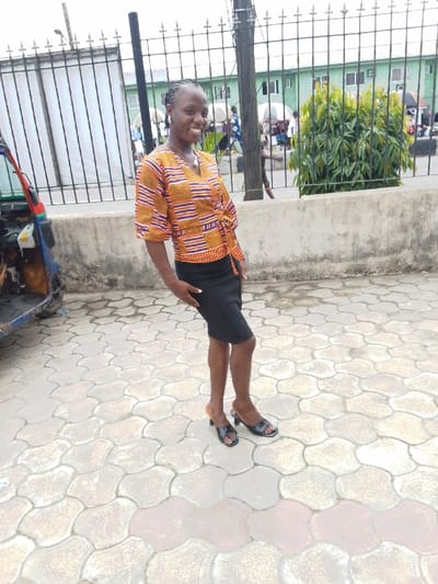 The image shows a young woman standing on a sidewalk in Yenagoa, Nigeria, at the location defined by the coordinates (latitude: 4.931, longitude: 6.268). The woman is smiling and wearing a colorful, striped blouse with a black skirt and black heeled sandals. She is standing in front of a fence with a green building behind it. The fence is made of metal and has a concrete base. The sidewalk is made of concrete blocks with an intricate pattern. There is a bush with green leaves on the right side of the image. The background is blurred.