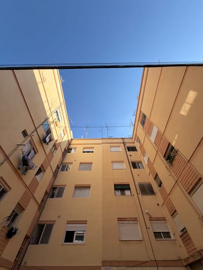The image shows an upward view of two apartment buildings facing each other in a narrow courtyard or alley in Castelló De La Plana, Spain. The buildings are painted in a light yellow ochre color with darker ochre horizontal bands. The sky above is a clear, vibrant blue. Several horizontal dark cables are stretched across the top of the frame, obscuring a portion of the sky.
The building on the left has several open windows. Laundry is hung from a clothesline extending from one of the windows, with various garments visible. Another window appears to have blinds partially open. Antennas are visible on the rooftop of the building on the right, along with some greenery in a window box.
The perspective is looking directly up, creating a sense of depth and emphasizing the height of the buildings. The lighting suggests it is daytime, with sunlight casting shadows on the building facades. The overall impression is one of a typical urban residential environment.