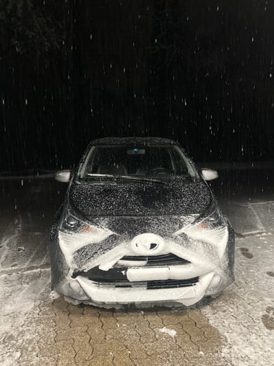 A black car is parked in a snowy environment, likely in Wetzikon (ZH), Switzerland, given the prompt context. The front of the car is heavily covered in snow, obscuring the grill and headlights. The windshield is visible but also covered in snow. The car is parked on a paved surface that is partially covered with snow and possibly ice. The background on the left side of the car is dark and appears to be unlit. The environment suggests a cold, possibly nighttime, winter scene with recent snowfall.