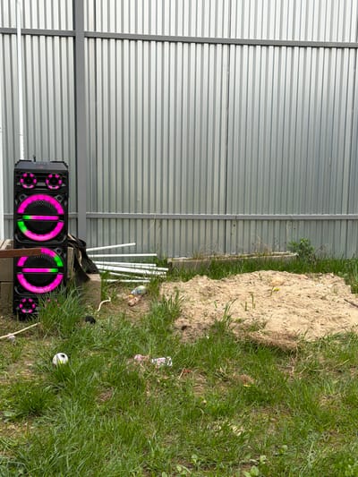 The image shows an outdoor scene with a corrugated metal fence in the background. To the left of the fence, there's a large black speaker with circular LED lights in pink and green. Some white PVC pipes are lying near the speaker and the fence. To the right, there is a mound of dirt and dense green grass. The location is likely Sverdlovskiy, Russia based on the user’s request. There are no people visible in the image. The overall impression is of an unkempt outdoor space next to a structure with a metal fence.
