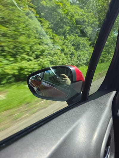 The image shows a view from inside a vehicle, looking out the passenger side window. The side mirror is visible, reflecting a hand holding a phone. The exterior environment is blurred due to motion, depicting lush green trees and foliage, indicative of a rural or park-like setting. The scene is captured during daylight. The location of Stockton-on-Tees, United Kingdom, is not discernible from the visual information in the image.