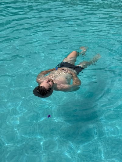 A person is floating on their back in a swimming pool. The water is clear and blue, with sunlight reflecting off the surface, creating shimmering patterns. The person appears to be relaxed, with their eyes closed and their arms and legs extended. They are wearing dark swim trunks. There is a small purple object floating in the water to the lower left of the frame. The bottom of the pool is visible, tiled in a mosaic pattern. The overall scene evokes a sense of peace and tranquility.