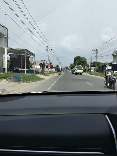 This media file captures a view from inside a vehicle, looking out at a road in Payameuneng, Indonesia. The scene is set on a paved road with some debris and vegetation along the side. Power lines are prominent in the sky, strung between utility poles.
Several objects and subjects are visible. On the left side of the road, there is a building with a covered outdoor area, a blue trash bin, and some vegetation. Further down the road, there are various vehicles, including a light blue truck carrying lumber, a black car, and several motorcycles. Two people are riding on one motorcycle in the foreground, a rider and a passenger, both wearing helmets. The rider has a light-colored jacket and the passenger appears to be wearing a dark jacket.
The sky is overcast with clouds, suggesting a day with diffused light, possibly morning or afternoon. The overall atmosphere appears to be typical of a roadside scene in a populated area.
Text is visible on a sign on the right side of the road, appearing to be a "76" logo and some Indonesian text. Another sign is visible further down on the left with some text that is not clearly legible.
The image provides a snapshot of daily life and traffic on a road in this Indonesian location.