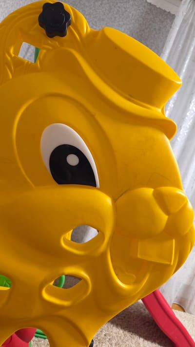 The image shows a close-up, eye-level view of a bright yellow plastic toy, likely part of a play structure. The toy is designed to resemble an animal's face, with a prominent white and black eye and visible front teeth. The surface of the toy shows some minor scuff marks and faint scratches. The background consists of a textured, light-colored surface, possibly carpet or a similar material, and a translucent white material, which could be a curtain or a sheet of plastic. A portion of a red slide is visible in the lower right corner of the image. The scene does not depict any individuals or indicate a specific location in Sannikovo, Russia, or any other city.