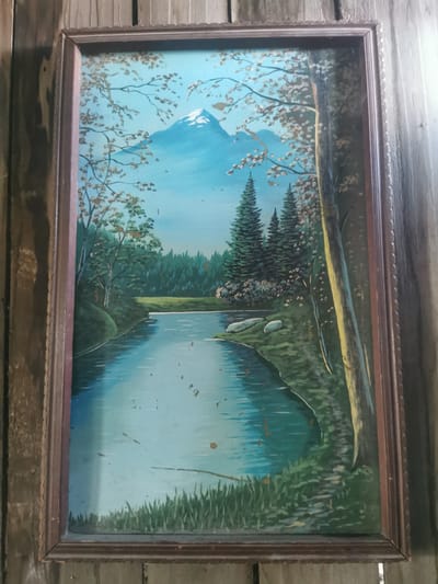 The image displays a framed painting of a natural landscape. The painting depicts a serene scene with a calm body of water flowing through a lush green environment. Tall, dark green evergreen trees dominate the background, forming a dense forest. In the distance, a majestic snow-capped mountain rises against a bright blue sky. The foreground features grass and the edge of the water. The painting is enclosed in a dark, ornate wooden frame. The overall impression is one of tranquility and natural beauty. The image itself is a photograph of this painting, and the background shows what appears to be wooden paneling, suggesting the painting is hanging on a wall. There is no information or visual indication within the image that links it to the city of Serov, Russia.