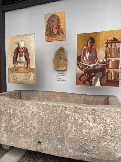 The image captures an indoor exhibition space, likely a museum or gallery, located in Drama, Greece. In the foreground, a large, weathered, rectangular stone sarcophagus or basin rests on a modern grey support on a dark, pebbled floor. Its exterior surfaces are intricately carved with faded inscriptions and relief motifs, including a distinct rosette-like pattern on its side.
The background wall, painted a plain light-grey, displays various artworks and artifacts. Three oil paintings are suspended from thin wires. On the left, a vertical painting depicts an older woman in a white chef's hat and apron, actively using a wooden rolling pin to flatten dough. Centered above, a horizontal portrait features a woman with long, curly brown hair, rendered with a contemplative expression. On the right, a vertical painting shows a young woman in a white sleeveless top seated at an ornate wooden desk, writing with a pen; a bookshelf filled with books is visible behind her.
Between the left and right paintings, a light-brown relief sculpture of a robed bust is mounted on the wall. Directly beneath this sculpture, two white rectangular informational plaques with black text are affixed to the wall. The ceiling structure above is dark.