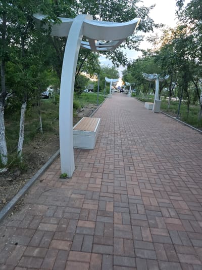 The image depicts a paved walkway in a park-like setting, likely in Satbayev, Kazakhstan. The walkway is constructed from reddish-brown rectangular pavers laid in a herringbone pattern. Along the left side of the walkway, there are modern, white, curved structures that appear to be canopies or decorative elements, providing shade. Several concrete benches with wooden slats are visible along the walkway, some with people seated on them. Trees with lush green foliage line the walkway and the surrounding area, suggesting a warm season. Sunlight filters through the trees, indicating it is likely late afternoon or early evening. The sky is visible in the background, with some light clouds. The overall scene conveys a sense of peacefulness and public recreation.