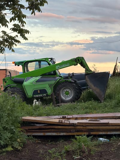 The image depicts a Merlo Turbofarmer telescopic handler parked outdoors near Cherni Vrah, Bulgaria. The machine is green and black. It has a large front bucket. The handler is parked on a grassy area beside metal gates. The sky is cloudy, with hints of sunset. A utility wire is visible above. Foliage is present in the foreground and background.