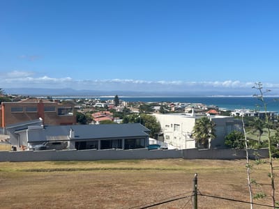 This image presents an elevated, panoramic view of a coastal town identified as Jeffrey's Bay, South Africa, under a clear blue sky.

The foreground consists of a large, undeveloped plot of dry, light-brown ground. A low, light-colored concrete or stucco wall with a horizontal top edge spans the mid-ground, separating this plot from the residential area below. To the right of the foreground, a tall, slender, leafless plant stalk with several upward-reaching branches is visible. Slightly left of center in the foreground, a weathered wooden utility pole stands vertically, with a small, dark bird perched on its apex; thin wires extend from the pole.

The mid-ground is dominated by a densely packed residential area featuring numerous houses with various architectural styles. Prominently in the center, directly behind the foreground wall, is a modern single-story house with a dark grey pitched roof, extensive dark-paned windows, and a white exterior; a cylindrical grey water tank is visible on its left side. To the left of this house, further down the slope, is a two-story brick building with multiple rectangular windows and a prominent chimney. To the right, another multi-story light-colored building with a flat roof is partially visible, surrounded by mature palm trees and green foliage. A turquoise-colored swimming pool is visible within a walled yard to the far left. Roofs are varied, including red tiles and various shades of grey.

The background features the Indian Ocean, displaying shades of deep blue and turquoise, with white surf visible along the coastline. A sandy beach separates the town from the ocean. Distant landmasses or sandbanks are visible further out in the bay. Beyond the ocean and town, a long range of hills or mountains stretches horizontally across the horizon, partially obscured by a band of low, white, wispy clouds. The sky above is uniformly bright blue, with minimal cloud cover. No people are visible within the frame.
