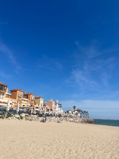 The image depicts a coastal scene in El Puerto de Santa María, Spain. On the right, a sandy beach occupies a significant portion of the frame. Adjacent to the beach is a series of buildings, mainly residential, constructed in a style typical of coastal Mediterranean architecture, featuring pastel colors and balconies. Palm trees are interspersed among the buildings. Between the buildings and the beach is a line of rocks, possibly serving as a breakwater or erosion control measure. In the background, there is a glimpse of the ocean horizon. The left side of the image is dominated by a clear blue sky with wispy clouds. The photo is taken from a low angle, enhancing the perception of height for both the buildings and the sky. The scene suggests a sunny day, with a calm atmosphere.