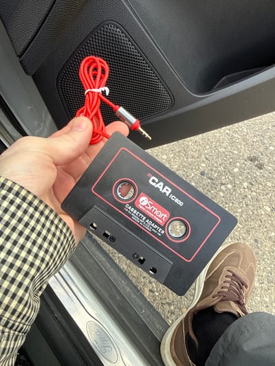 A person is holding a black cassette adapter with red trim, a brand name "mCAR" and "Smart" logo are visible, along with text that reads "CASSETTE ADAPTER" and "MADE IN CHINA". A red audio cable with a silver and red jack is attached to the adapter and coiled. The person is wearing a checkered jacket and dark pants. A brown shoe and a black sock are visible. The background shows the interior of a car door with a speaker grate and a door pocket. The floor outside the car is visible, which appears to be concrete with some gravel. A silver door sill with the "Ford" logo is also visible. The scene is outdoors. The location is Mayrtup, Russia.