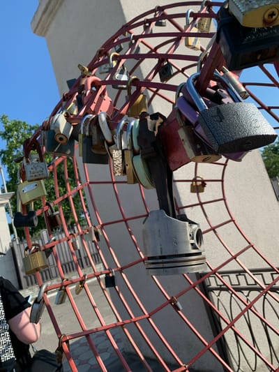 In the city of Korzhevo, Moldova, a bright red metal structure resembling a curved fence is adorned with numerous padlocks of various sizes, colors, and conditions. Some padlocks are shiny and new, while others are rusted and weathered, suggesting they have been in place for some time. The structure appears to be a decorative art piece or monument. Suspended from the structure is a metallic component, possibly a piston from an engine, further adding to the unconventional nature of the display. In the background, a light-colored building and a clear blue sky are visible. On the left side of the frame, a person is partially visible, seated, and appears to be interacting with a smartphone. The lighting suggests it is a sunny day.