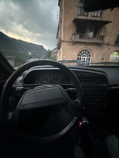The image, taken from the interior of a car in Tumanyan, Armenia, captures a view of the city's mountainous surroundings and architecture. The foreground is dominated by the steering wheel and dashboard of the car, with visible instruments including a speedometer and tachometer. A manual gear shift is also prominent. In the background, a multi-story building with balconies and arched windows is visible, suggesting an older architectural style. Beyond the building, steep, rugged mountains rise under a cloudy sky, with glimpses of the sky tinged with sunlight peeking through the clouds. The overall scene depicts a moment of stillness within a car, looking out onto the unique landscape of Tumanyan.