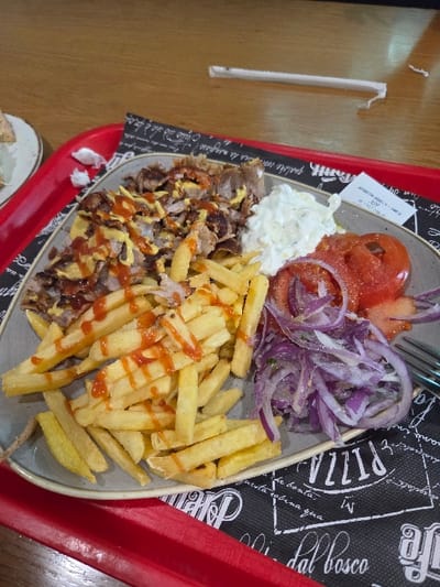 A prepared meal is depicted on a light grey ceramic plate, centrally positioned on a red plastic tray and a black paper placemat with white "PIZZA" and "dal bosco" text. The plate contains a portion of sliced gyros meat, topped with yellow and red sauces. Alongside the meat is a substantial serving of golden-brown french fries, drizzled with red sauce. To the right, a scoop of white creamy sauce (likely tzatziki) is present, next to several slices of red tomato and a mound of finely sliced red onions. A silver-colored fork is partially visible on the right edge of the plate. A small white slip of paper, tucked under the plate on the right, identifies the establishment as Aigaion Gyros (Αιγαίον Γύρος) located at Eth. Antistasis 14, Polykastro 612 00, Greece. The background consists of a light brown wooden table surface, on which a crumpled white straw is visible. A portion of a second plate is present on the far left. No individuals are depicted. The scene suggests an indoor dining environment.