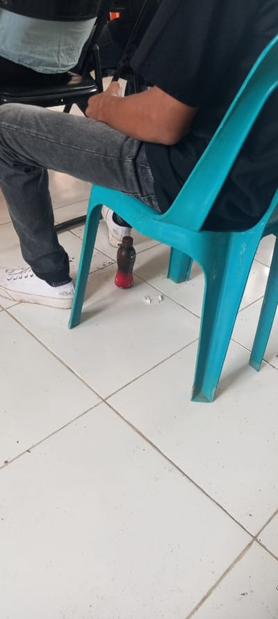 A person wearing gray jeans and white sneakers sits on a blue plastic chair. The person is wearing a black shirt. A chocolate drink bottle sits on the white tiled floor beneath the chair. There are some pieces of trash next to the bottle.
