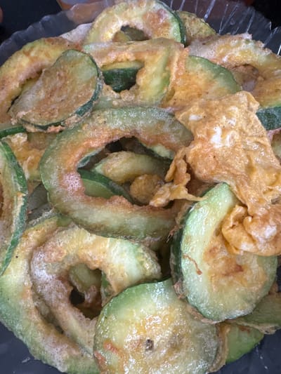 This is a close-up, high-angle shot of a pile of fried zucchini slices in a clear bowl. The zucchini slices are round and have a light green color with some brown, crispy edges. They appear to be battered and fried, with some pieces coated in a yellowish batter. There are also pieces of what appears to be scrambled egg mixed in with the zucchini. The lighting is bright, and the focus is on the food, suggesting it is a still life photograph. The setting is an indoor environment, possibly a kitchen or dining area, with a dark background. There are no people or any indication of a specific location in the city of Batken, Kyrgyzstan.