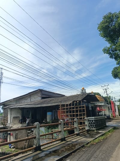 An eye-level, sunlit shot showcases a low-angle perspective of a scene. A weathered building with a corrugated roof sits adjacent to a bridge with metal and concrete railings. Overhead, a network of power lines crisscrosses the expansive sky. In the background, a mosque with golden domes is visible beyond the building. A nearby utility pole and trees add to the urban landscape. The scene is characterized by a mix of infrastructure, architecture, and nature.