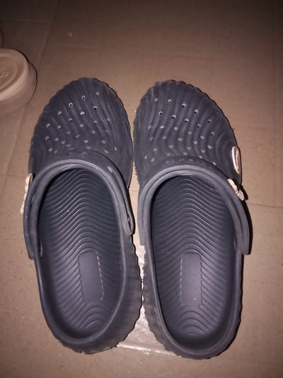 In Bida, Nigeria, a pair of dark grey clogs are placed side-by-side on a tiled floor. The clogs are made of a textured rubber material with a series of circular perforations on the upper part. A strap is visible on the side of each clog, with a small, light-colored button-like adornment attached to it. The floor appears to be made of flat, square tiles with a light brown hue and some scattered dark speckles. The lighting in the scene is dim, casting shadows around the clogs. To the left of the clogs, a portion of a round, light-colored object, possibly a container lid, is visible. The perspective of the image is from above, looking down at the clogs.