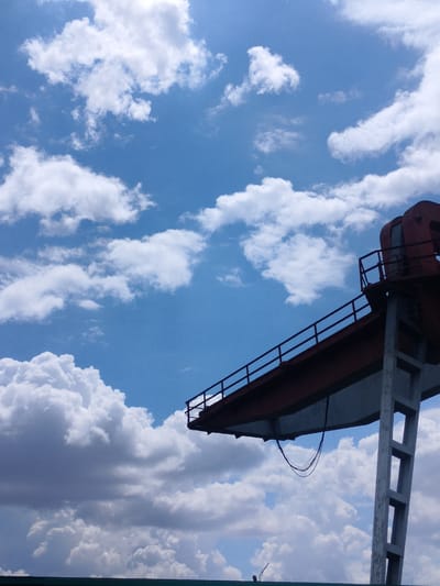 This is a low-angle, full-shot, outdoor photograph taken against a bright blue sky filled with fluffy white clouds. The clouds vary in size and density, with some appearing larger and more shadowed, suggesting depth and possibly an approaching weather system.
Dominating the right side of the frame is the upper section of a large, industrial crane. The crane is painted a deep red and appears to be constructed of metal beams and supports. A narrow walkway with a dark railing runs along the length of the horizontal boom of the crane. The vertical support structure, made of grey concrete with rectangular openings, is also visible. Electrical wires hang loosely from the underside of the crane's boom.
The foreground shows the top edge of a dark green structure, possibly a building or another part of the industrial infrastructure, which is out of focus. In the far distance, a small, silhouetted figure can be seen standing on this structure, and a thin, dark pole is also visible.
The overall impression is one of a bright, possibly daytime scene, given the clear blue sky and bright lighting. The presence of the industrial crane suggests an industrial area or a port. The weather appears to be fair, with scattered clouds. No text is visible in the image. The scene evokes a sense of scale and the grandeur of industrial machinery against the vastness of the sky.