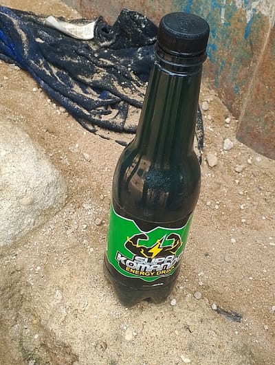 The image shows a bottle of "Supa Komando" energy drink lying on the ground. The bottle is dark green and features a green label with a muscular arm holding a lightning bolt, with the words "Supa Komando Energy Drink" printed in white and yellow. The bottle is partially obscured by a dark blue cloth lying nearby. The background is a rough, sandy surface, possibly a construction site or an area with loose aggregate. The coordinates 5.935 latitude, 5.67 longitude, place the image in the city of Ogharefe, Nigeria.