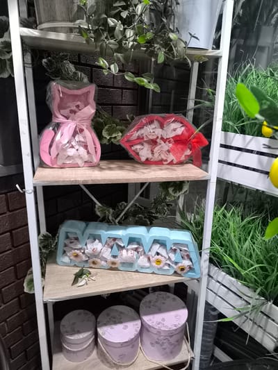 This media file showcases a retail display of gift-wrapped chocolates and decorative boxes, likely in a store or market stall in Vulcănești, Moldova. The items are arranged on white shelving units.
On the top shelf, a pink, cat-shaped gift box filled with chocolates, tied with a pink ribbon, is visible. To its right, a red heart-shaped container, also filled with chocolates and adorned with a red ribbon, is present. Surrounding these items are various green plants, adding a decorative touch.
The middle shelf holds a blue tray filled with chocolates and small decorative flowers. A single green leaf rests on the shelf in front of the tray.
The bottom shelf displays two round decorative gift boxes with floral patterns in shades of pink and lilac, tied with rope.
In the background, to the right, a white wooden planter box filled with lush green grass and a hint of yellow fruit (possibly lemons) can be seen, suggesting an outdoor or garden-themed display. The overall setting appears to be indoors, with some elements hinting at an outdoor or natural aesthetic. There is no indication of people or a specific time of day or weather conditions.
Visible text on the chocolate wrappers reads "Raffaello".