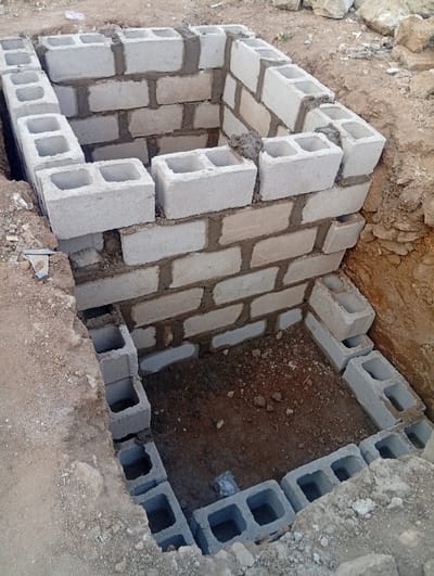 A partially constructed rectangular structure made of concrete blocks sits within a dug-out pit. The foundation layer, consisting of blocks laid in a rectangular pattern, is partially filled with dirt. The walls rise from the base, constructed with additional concrete blocks held together by mortar. The wall's top edge is uneven, indicating ongoing construction. The surrounding ground is a mix of dirt and rocks, with evidence of excavation visible.