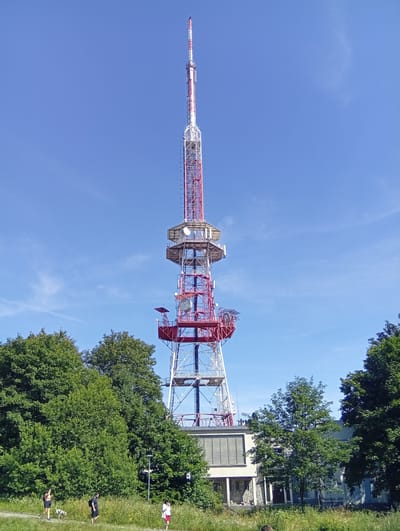 This is a full-frame, eye-level, slightly low-angle shot of the Radio Mast in Sobótka, Poland, on a clear, sunny day with a vibrant blue sky. The mast, a prominent telecommunications tower, dominates the upper half of the frame. It is a lattice structure, painted in sections of white and bright red. Various antennas and dishes are visible attached to its various levels, along with what appear to be observation platforms or equipment housings.
In the foreground, a lush green area is filled with trees and tall grass, interspersed with wildflowers. A path or walkway runs horizontally across the lower portion of the image, with several individuals present. On the left side of the path, a woman in athletic wear with a dog is walking, and to her right, a man wearing a black t-shirt and shorts is also walking. Further to the right, a person in red shorts and a white t-shirt is walking away from the camera. A lamp post is visible behind the walking individuals.
Behind the trees and walkway, a low-rise building with a distinct grid-like pattern on its upper section is partially visible. The building's facade is light-colored, with visible windows and doorways. The overall impression is of a sunny day in a park-like setting, dominated by the imposing structure of the radio mast.