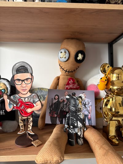The image displays a shelf filled with various decorative items, none of which indicate a specific location in Малое Седельниково, Russia. On the shelf, there is a caricature-style cutout of a person holding a red electric guitar, wearing a gray baseball cap and glasses. Behind it, a large burlap doll with button eyes and stitched features is positioned centrally. To the right of the doll are two gold-colored bear-shaped figures and a small crocheted yellow creature. In front of the doll, a framed photograph of a group of people is visible, along with a black and white splattered toy figure. The shelf itself is made of wood.