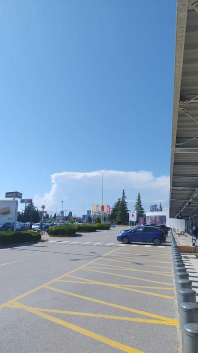 The image shows a bright, sunny day with a clear blue sky. In the foreground, a parking lot is visible with yellow lines painted on the asphalt. A blue car is parked on the right side of the lot, next to a row of bollards. Beyond the parking lot, there are several buildings with signage, including an IKEA store. There are also trees and some people walking in the distance. The scene appears to be a commercial area. The location is Pylaia Municipal Unit, Greece.