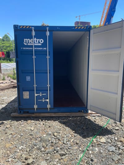An open blue shipping container sits on a wooden pallet on a gravel lot. The container is branded with "metro LOGISTICS" and "1st GEORGIAN CONTAINER LINE." The container doors are open, revealing the interior. In the background, laundry hangs on a clothesline. A construction crane and an unfinished building are visible further in the distance. The scene appears to be outdoors in a somewhat industrial or construction area. The location is identified as Chakvi, Georgia.