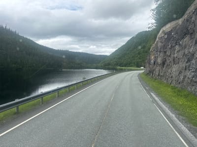 The image shows a two-lane asphalt road curving to the right alongside a dark body of water. The road has a white line marking the edge and a broken yellow line dividing the lanes. A metal guardrail separates the road from the water. The water is bordered by a steep, tree-covered mountain slope on the left. On the right side of the road, a sheer rock face rises, with some vegetation growing at its base and top. The sky is overcast with grey clouds. In the distance, a small white vehicle can be seen on the road. The scene is consistent with a road in a mountainous, coastal region, potentially near Mosjøen, Norway, given the landscape and the presence of a fjord-like body of water.