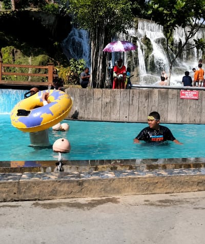 The image depicts a lively water park scene at a location in Silaiyajae, Indonesia, featuring a cascading waterfall as a backdrop. In the foreground, a young boy wearing goggles and a black t-shirt is swimming in a bright blue pool. To the left, a large yellow and purple inflatable ring floats in the water, with a child's feet visible inside. Several other people are visible around the pool area, with some sitting near the edge and others standing closer to the waterfall. A pink and white umbrella provides shade for a person seated in a red chair. Wooden fencing and a concrete wall form the perimeter of the pool area. A sign in red text is attached to the wooden wall. The environment is sunny and appears to be a popular recreational spot.