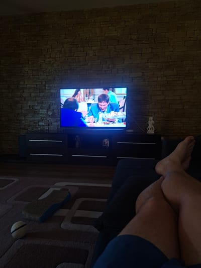 The image depicts a living room scene in Dumbrăvița, Romania, characterized by a dark, modern aesthetic. A large flat-screen television displays a scene of people dining at a table, with one individual in a blue suit prominently featured. The television is placed on a dark, minimalist entertainment unit with several drawers. To the right of the television, a small, abstract white sculpture is visible. The wall behind the television and sculpture is covered in textured, dark stone veneer. In the foreground, a person's legs and feet are visible, suggesting they are relaxed on a dark sofa, facing the television. On the floor, to the left, a cat scratching post with a blue base and a textured surface is present, along with a round, woven ball. The lighting in the room appears to be dim, with the primary illumination coming from the television screen. The flooring is a patterned carpet.