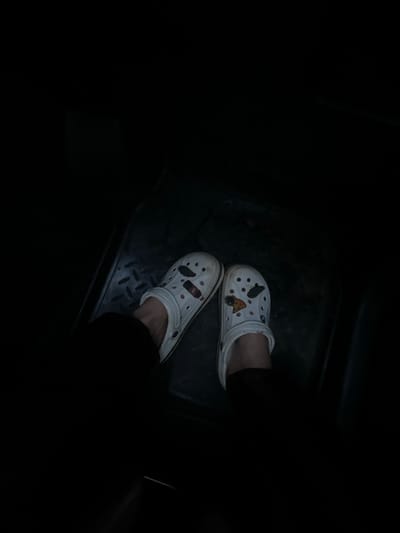 The image shows a person's feet wearing white Crocs with various charms, resting on a dark, textured surface, possibly a floor mat. The scene is dimly lit, with the majority of the image in shadow. The Crocs have charms depicting items like a lighter and a slice of pizza. The background is dark and out of focus, making it difficult to ascertain the specific environment. There is no information in the image to suggest the location is in the city of Meshcheryaki, Russia.