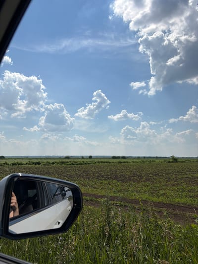 The image captures a view from a car, looking out the side window. In the foreground, a side-view mirror reflects a white car and a person's arm with a visible thumb. The mirror is framed by the car's interior and the exterior view is dominated by a vast, cultivated field stretching towards the horizon. The field is a mix of dark soil and emerging green crops. Beyond the field, a line of trees and distant structures, possibly industrial, are visible against a bright blue sky filled with scattered white clouds. The scene appears to be in a rural area, as suggested by the expansive farmland and the absence of dense urban development.